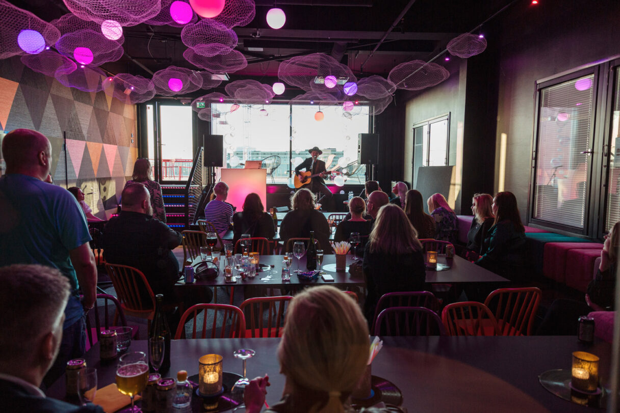 Circuksen edustustila iltatilaisuudessa, jossa ihmiset istuvat pöytien ääressä tunnelmavalaistussa salissa ja lavalla esiintyy bändi
