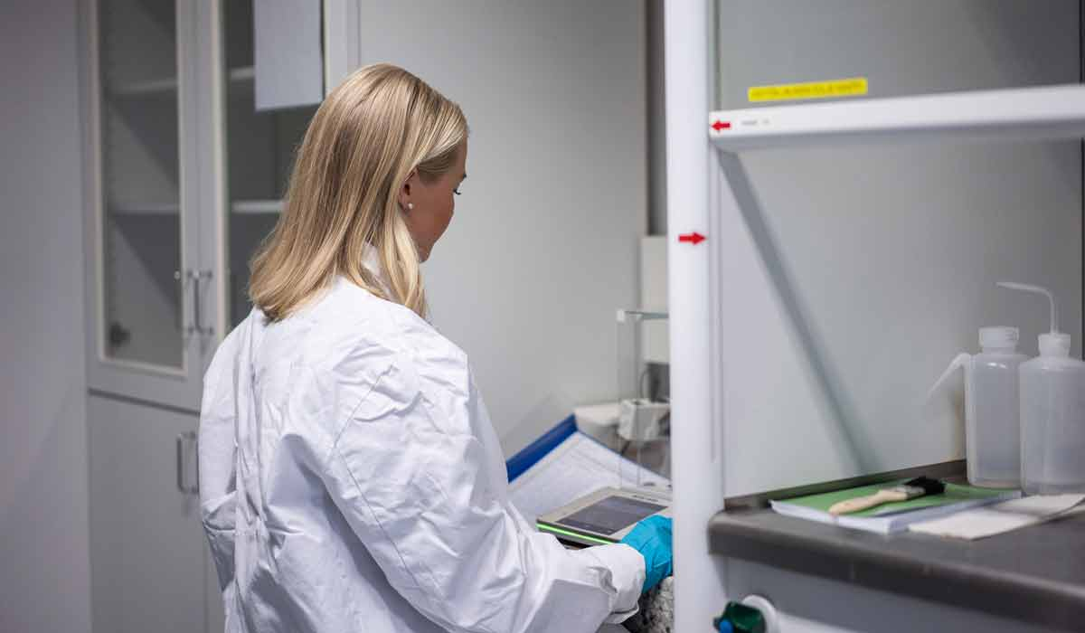 Henkilö työskentelee laboratoriotilassa käyttäen suojatakkia ja käsineitä tutkimuslaitteiden äärellä.