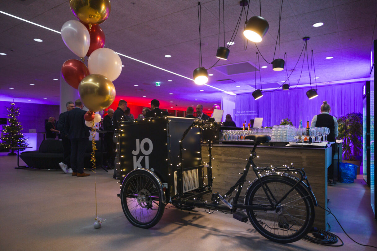 Joki‑brändätty tarjoilufillari koristeltuna valoilla ja ilmapalloilla tapahtumatilassa.