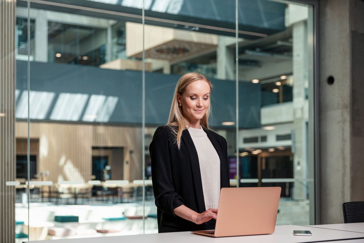 Vaaleahiuksinen nainen seisoo työpöydän ääressä ja käyttää tietokonetta