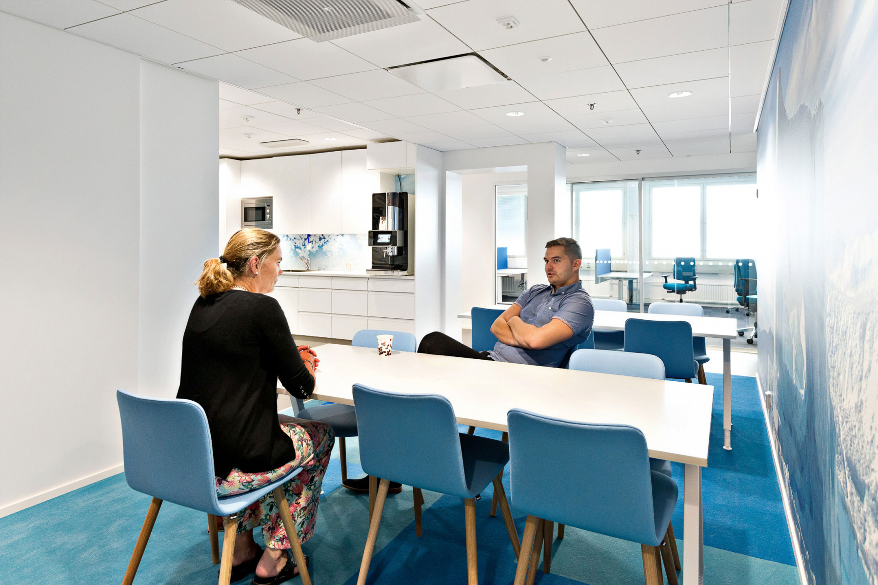 Kaksi henkilöä istuu keskustelemassa Werstaan valoisassa taukotilassa, jossa on keittiö ja pitkä pöytä sinisine tuoleineen.