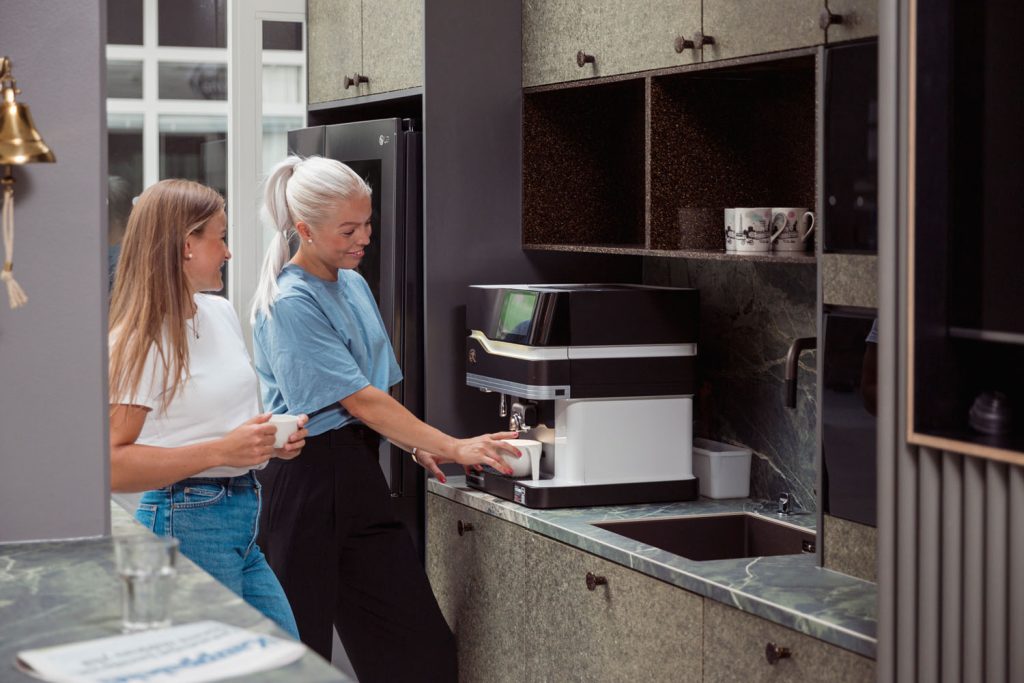 Kaksi ihmistä ottamassa kahvia kahvikoneesta vihreän sävyisessä toimiston kahviossa.