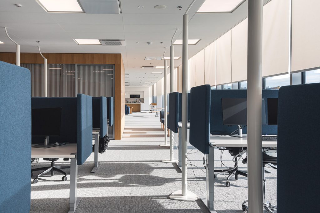 Rivissä sijaitsevia työpisteitä avoimessa toimistotilassa, joissa on työtuolit, näytöt ja väliseinäkkeet luonnonvalon valaisemassa ympäristössä