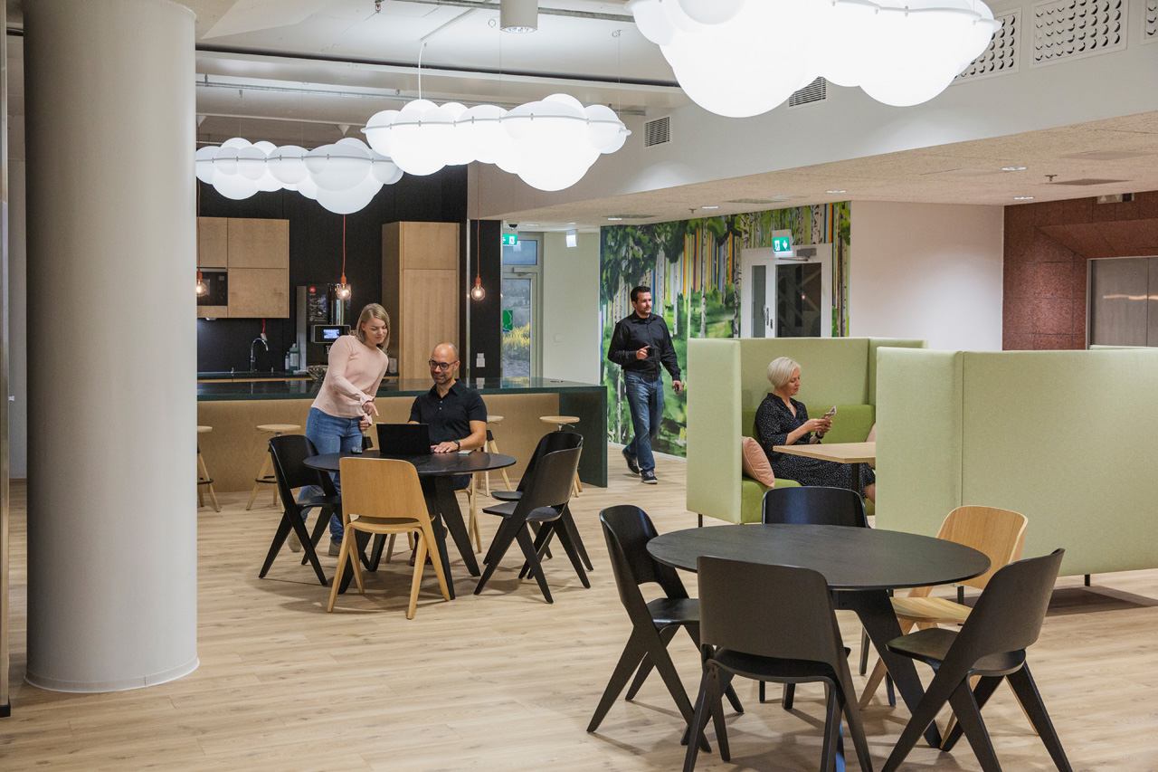 Ihmiset työskentelevät ja keskustelevat modernissa coworking‑tilassa, jossa on pilvimuotoiset valaisimet, keittiötausta ja vihreitä työskentelykoppeja.