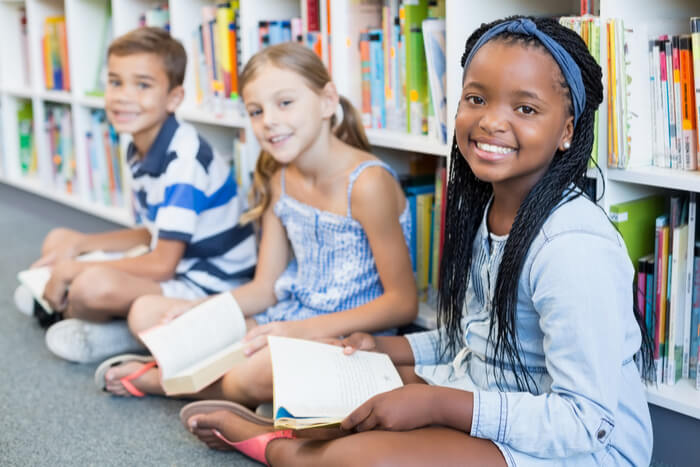 Portrait of smiling school kids sitting on floor and reading book in library