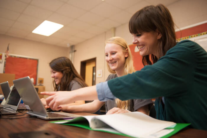 three students seated at their desks with their laptops and one of them pointing at the screen