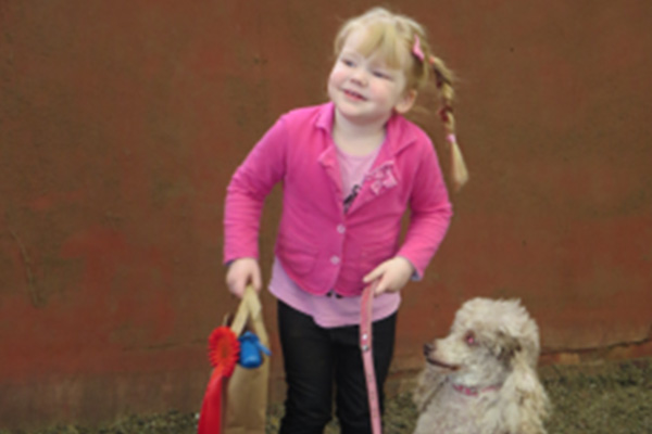 A girl wearing a pink top and black leggings holding a pink lead with a little white poodle looking up at her.