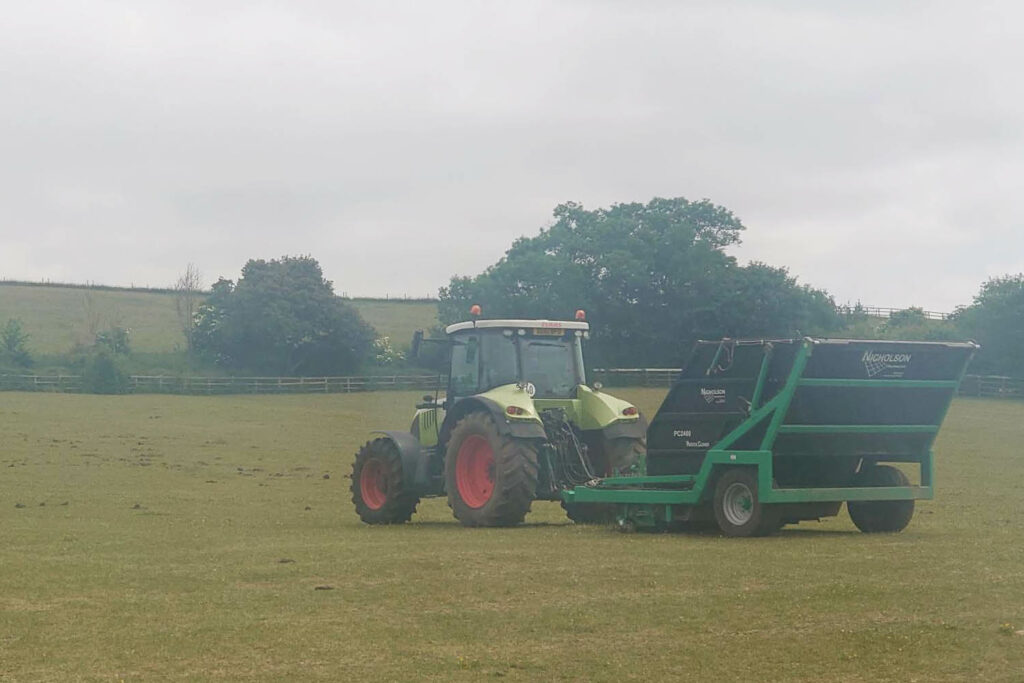 Large green tractor towing a paddock cleaner collecting horse droppings in a large field with a wooden fence and trees in the background