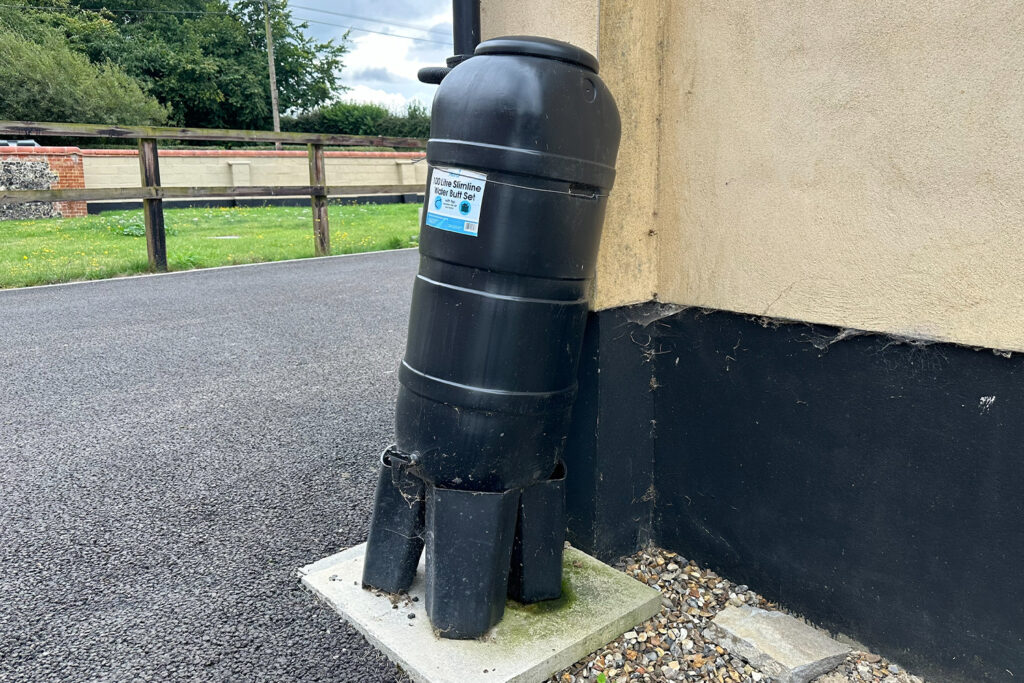 Dark green water butt on a dark green stand next to a cream and black wall attached to a black drain pipe