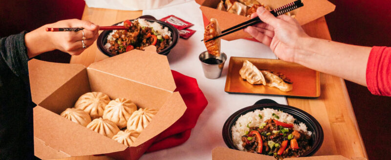 A table spread of Wow Bao food items. Boxes of bao, rice bowls, and potstickers. There are 2 hands with chopsticks, one is dipping a potsticker in sauce, the other is reaching for a rice bowl.