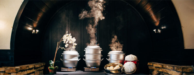 3 metal steamer baskets with steam coming out of them sitting on top of a table with a black table cloth. There is a steamer ring showing freshly steamed bao. The table has flowers to the left of it, and the table is sitting beneath a dark wood archway with brick accents.