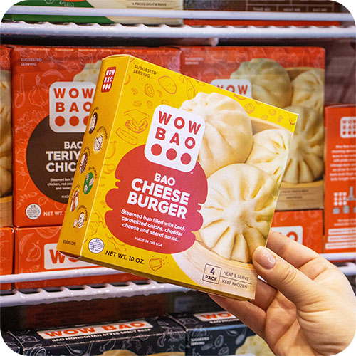 A hand holding a bright yellow box of Cheeseburger bao,  with other flavors of bao in a freezer case in the background.