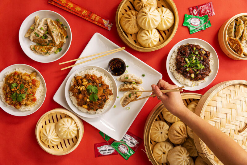 A large table spread of bao in large and small steamer baskets, potstickers and dumplings, and rice bowls. There are chopsticks and soy sauce packets scattered on the red table cloth.