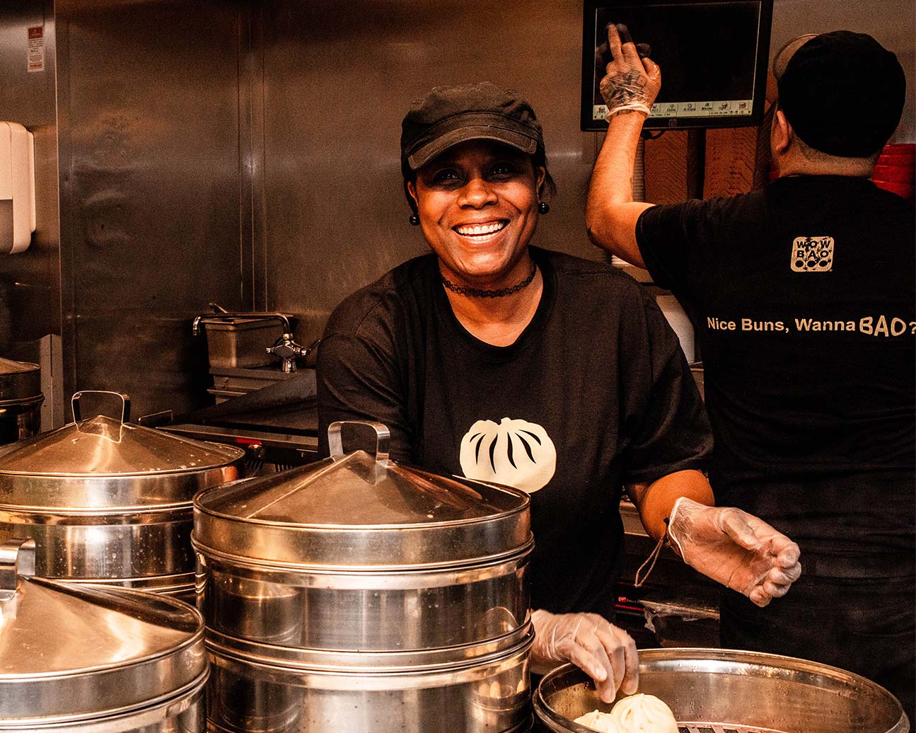 A Wow Bao team member, wearing a Wow Bao shirt, smiling towards the camera in front of metal steamers.