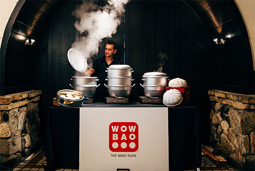 3 metal steamer baskets with steam coming out of them sitting on top of a table with a black table cloth. A person is standing behind the table lifting the lid off of a steamer basket. There is a steamer ring showing freshly steamed bao. The table has flowers to the left of it, and the table is sitting beneath a dark wood archway with brick accents.