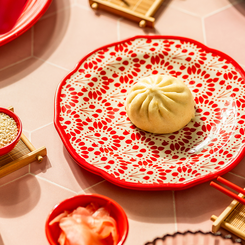 A steamed bao on a red patterned plate on a pink tile background