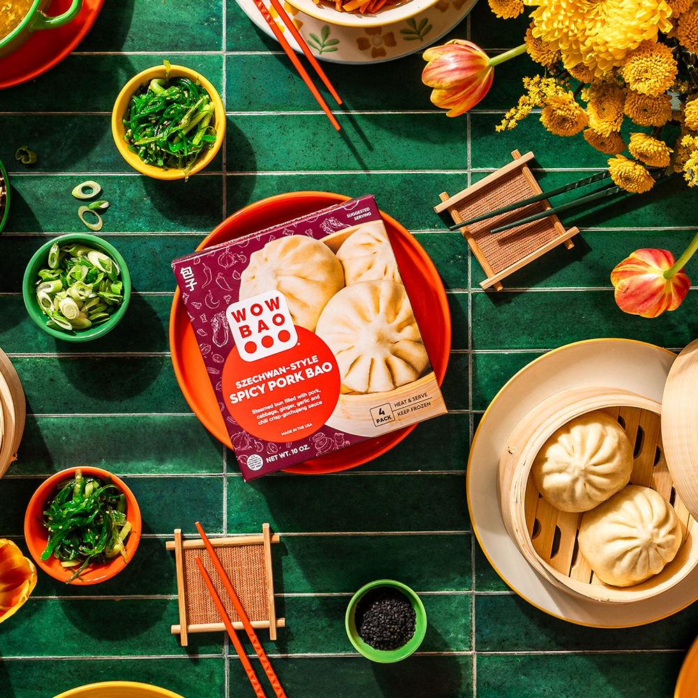 A box of Szechwan Style Spicy Pork Bao next to plates of freshly steamed bao on a green tile background with chopsticks, scallions, peanuts.