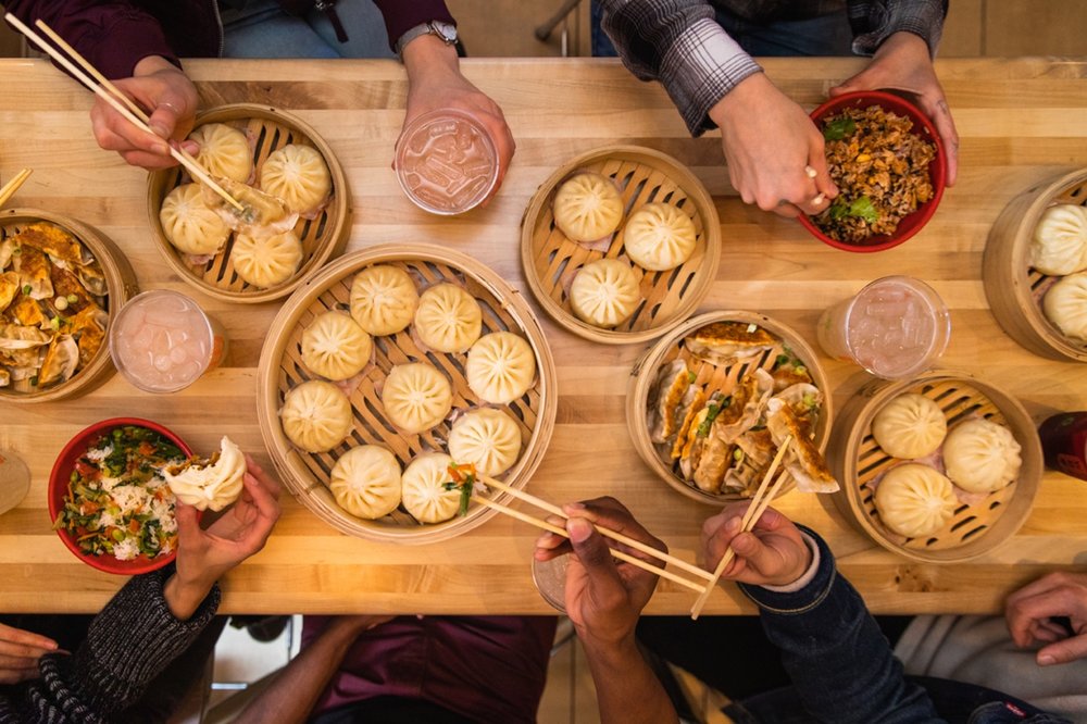 An overhead shot of a long wooden table of food. There are big and small steamer baskets full of bao, potstickers, and rice bowls. People are sitting at the table, their hands reaching for the food.