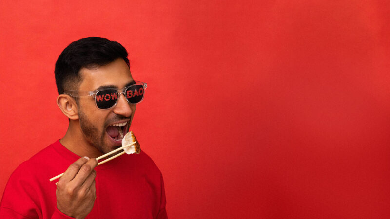 A person with Wow Bao sunglasses on, with chopsticks in their hand holding a potsticker, about to take a bite