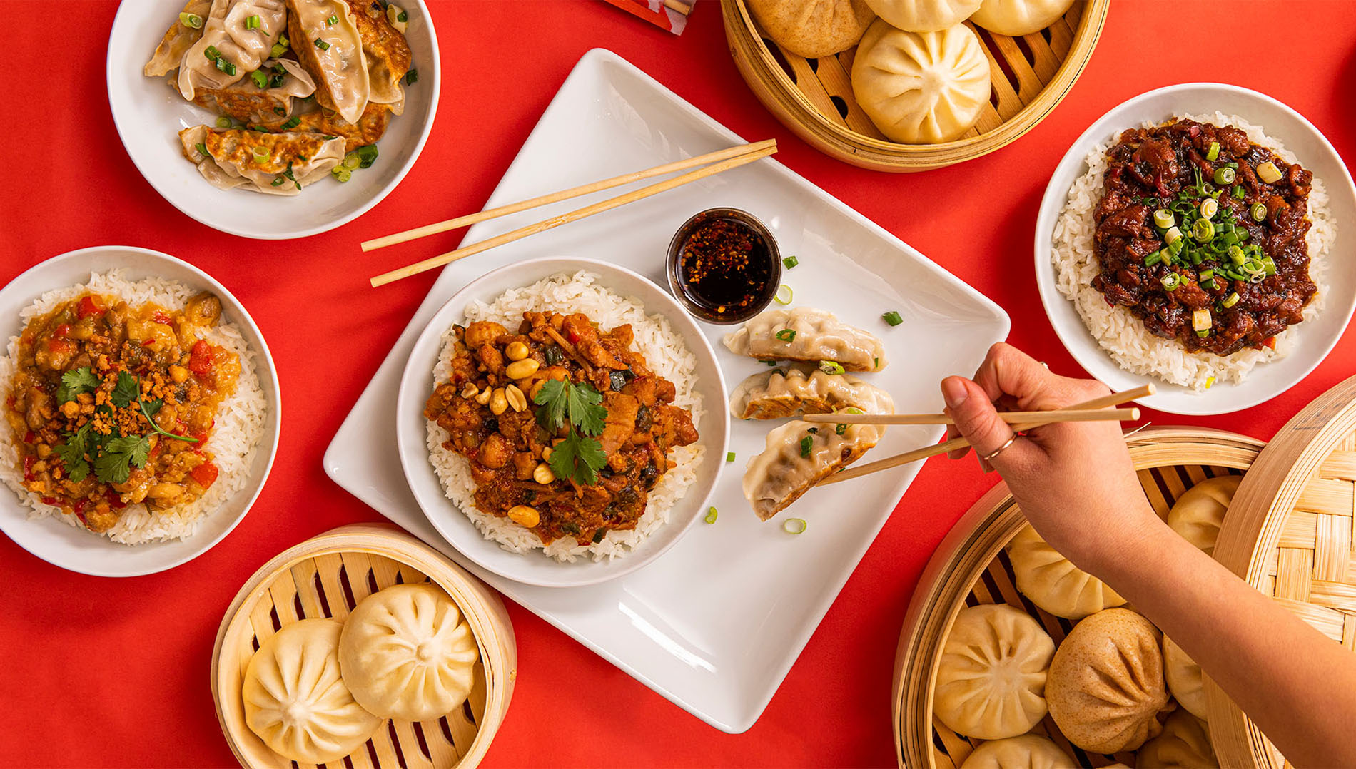 overhead photo of a table full of food including fried rice, bao and a hand holding chopsticks grabbing a pot sticker