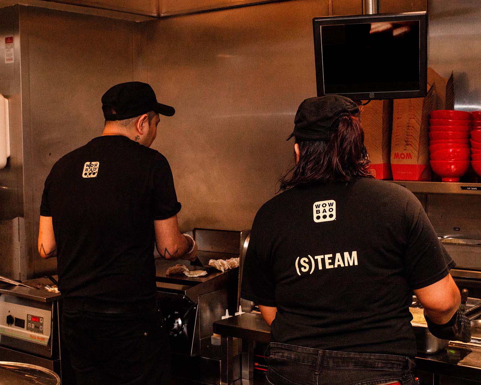 Two team members wearing Wow Bao shirts, one is cooking potstickers, while the other is making a rice bowl.