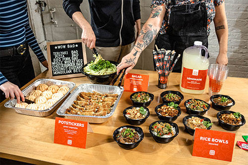 A table full of Wow Bao Catering, showing our bao, potstickers, and rice bowl that Wow Bao can cater for you. 3 people are reaching for food.