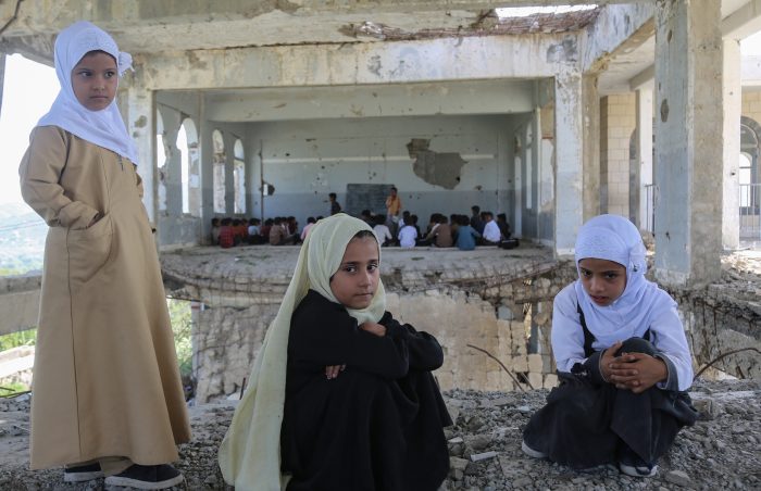 3 Yemeni girls in the destroyed school hall with a class taking place in the background.