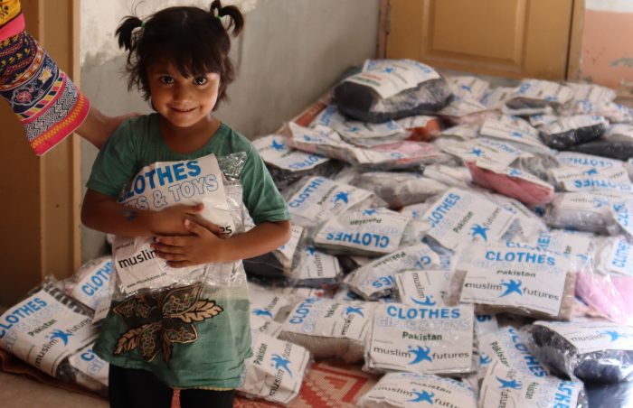 A young girl in Pakistan holds onto a packet of good quality donated clothes, with a pile of donated clothes in packets in the background.