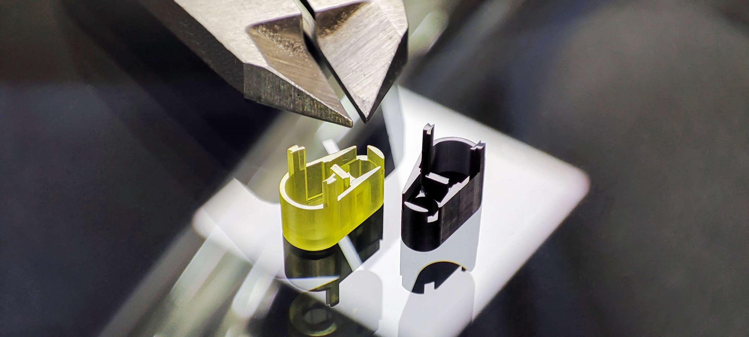 Two small 3D-printed parts, one yellow and one black, placed on a reflective surface with the tip of a pair of pliers above them for scale.