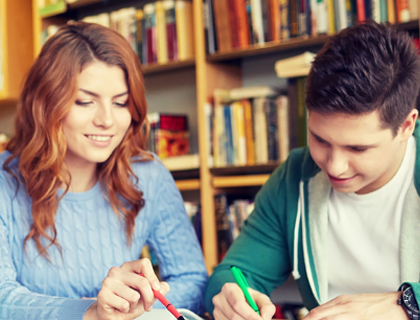 Dos estudiantes sentados en una biblioteca, escribiendo y estudiando juntos con libros al fondo.