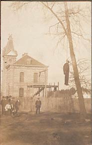 Unidentified corpse of African American male. Gallows, courthouse-jail, and windmill in background. Nine onlookers, two young boys. 1900-1915. Location unknown.