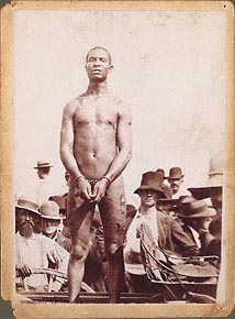 African American male standing on buggy, facing camera, stripped, deep lacerations and wounds, his handcuffed hands placed to cover his genitals. lynch mob. Circa 1900, location unknown.
