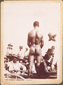 African American male standing on buggy, back to the camera, feet shackled, stripped, deep lacerations and wounds. Onlookers, one with a bloodstained shirt. Circa 1900, location unknown.