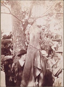 Lynching of African American male, victim hanging in a tree, stripped, lower body wrapped in coarse blanket. Lynch mob. Circa 1900, location unknown.