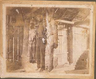 The corpses of Ernest Harrison, Sam Reed, and Frank Howard hanging from a rafter in a sawmill, jagged circular blade in lower right hand corner. September 11, 1911, Wickliffe, Kentucky.