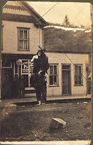 Unidentified corpse of badly beaten white male in shredded clothes hanging from rope stretched over unpaved street, onlookers in background. Circa 1900, Virginia City, Montana.
