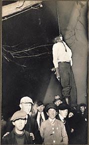Lynching of gagged black male hanging from a tree in autumn. Large crowd of onlookers, mostly boys. Circa 1920, location unknown.
