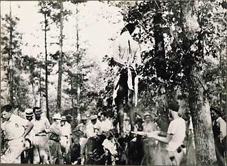 The lynching of W. C. or R. C. Williams, his body hanging from oak tree, lower body covered with kitchen apron, blood streaming down legs suggests castration, onlookers include white men and young children.
