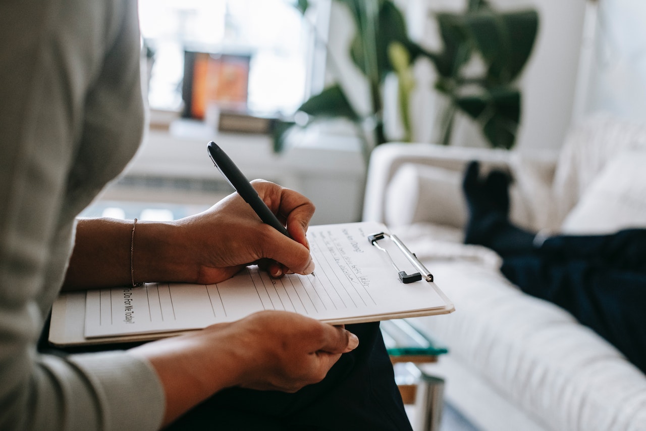 A therapist write information on a clipboard while a client lies on a couch
