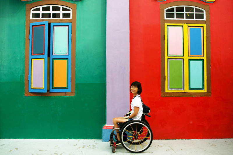 A woman in a wheelchair sits sideways smiling at the camera in front of a bright colored wall.