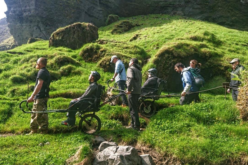 A group of friends travers uneven ands using a joëlette wheelchair, one of them sits on it, and two of them carry the chair working as a team.