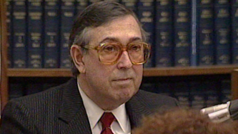 A man in glasses testifies in front of a bookshelf.