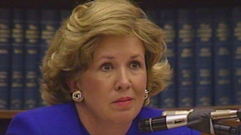 A woman testifies in front of a bookshelf.