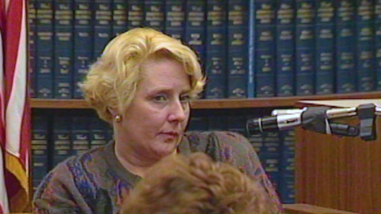 Betty Broderick sits in front of a bookshelf of blue books.