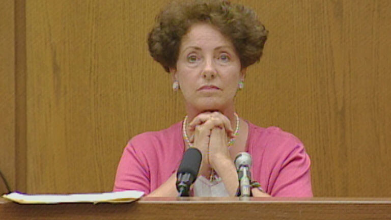 a woman holds her hands folded under her chin on the witness stand