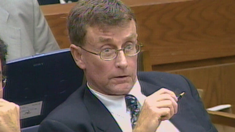 A man in a suit sits in court holding a pen