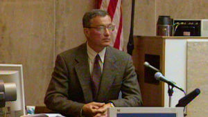 A man in a suit sits in front of a microphone in court