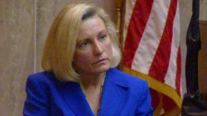 A woman in a blue blazer sits in front of an American flag