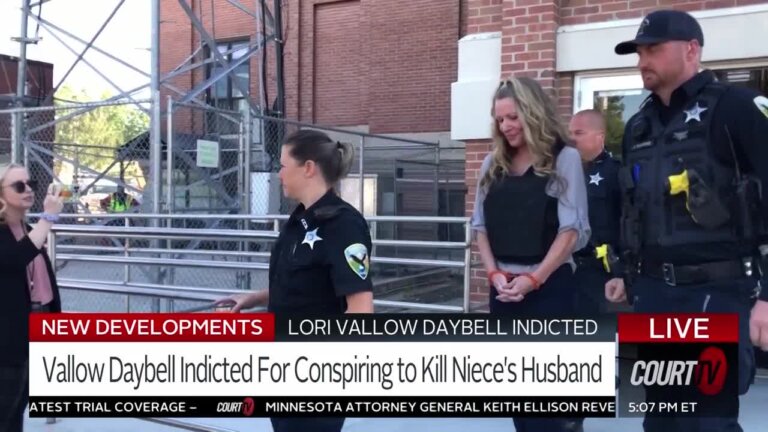 Lori Vallow Daybell, flanked by police, walks while wearing a bullet-proof vest