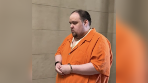 A man stands in an orange jail jumpsuit and handcuffs